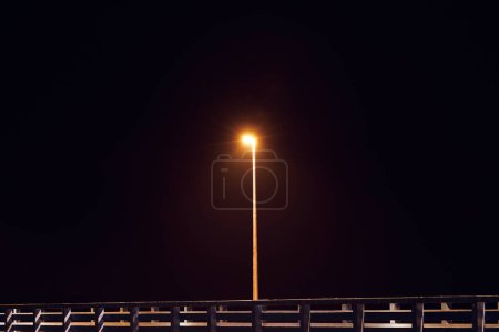 Einsamer Laternenpfahl wirft schummriges warmes gelbes Leuchten entlang der Landungsbrücke, die vor pechschwarzer Leinwand des Nachthimmels steht, zeichnet ein poetisches Bild der Seebrücke in der Stille der Nacht, der Gelassenheit einsamer Momente