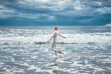 Photo for Happy hairless girl with alopecia in white futuristic suit dancing on beach bathed by ocean waves, performance of bald strong female artist, overcoming challenges of life and self confidence - Royalty Free Image