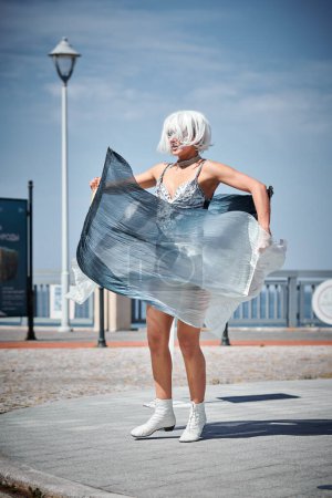 Young sexy girl in space silver micro skirt dancing with performance fan waving gracefully, female outdoor dance on seaside promenade creating outdoor spectacle harmonizes with rhythm of sea waves