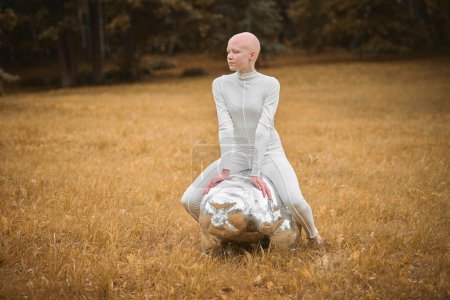 Photo for Young hairless girl with alopecia in white cloth sits on tardigrade figure in fall park, surreal scene with bald teenage girl confidently embraces her unique beauty - Royalty Free Image