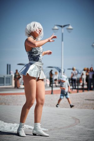 Foto de Joven chica sexy en el espacio micro falda de plata bailando con movimientos suaves, femeninos y elegantes, actuación de danza al aire libre femenina en el paseo marítimo creando un espectáculo al aire libre excitante - Imagen libre de derechos