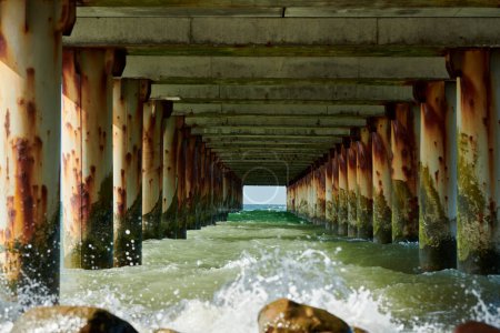 Rostige Säulen der alten Seebrücke schaffen eine faszinierende Meereslandschaft mit Blick unter der Seebrücke, unruhige Meereswellen, die auf Felsen krachen und eine harmonische und doch kraftvolle Küstensymphonie symbolisieren