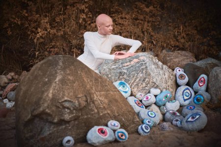 Young hairless girl with alopecia in white futuristic costume lying pensively among surreal landscape with many eyes stones, symbolizes introspection and reevaluation of individuality