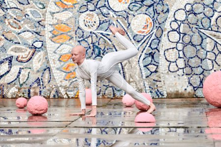 Tanz der jungen Ballerina mit Alopezie im weißen futuristischen Anzug mit plastischen und flexiblen Bewegungen zwischen rosa Kugeln auf abstraktem sowjetischen Mosaik, symbolisiert den Selbstausdruck