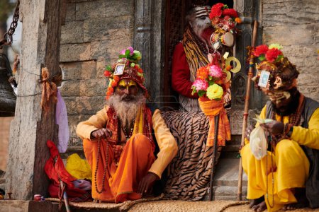 Foto de Katmandú, Nepal - 01.02.2023 - Tres Sadhu vestidos con ropa de color azafrán se sientan en el antiguo santuario votivo de Pandra Shivalaya en el templo de Pashupatinath, ascético Sadhu orando y rogando limosna - Imagen libre de derechos