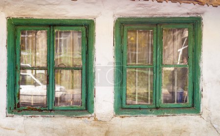 Foto de Dos viejos marcos de ventanas de madera verdes en una pared blanca y erosionada. La pintura está astillando y el vidrio refleja la escena exterior. - Imagen libre de derechos