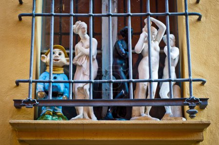 Foto de Una variedad de estatuas, incluyendo un colorido personaje de dibujos animados y figuras humanas clásicas, se muestran detrás de las barras de metal en una repisa de la ventana contra una pared naranja. - Imagen libre de derechos