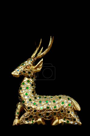 A detailed golden deer statue adorned with green and blue gemstones, captured against a black background. The craftsmanship highlights intricate designs and patterns