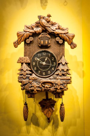 Eine sorgfältig geschnitzte hölzerne Kuckucksuhr vor leuchtend gelbem Hintergrund. Die Uhr zeigt oben einen Vogel und Blätter, unten Pendel und Tannenzapfengewichte. Eine zeitlose Zurschaustellung von Handwerkskunst.