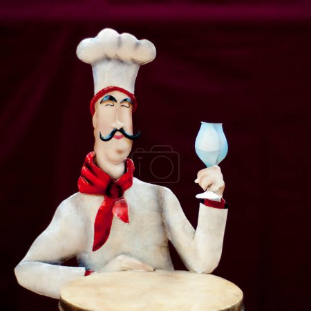 Foto de La figura del chef, hecha de un material pálido, lleva un sombrero blanco y una bufanda roja. Sostiene una copa de vino clara y se sienta al lado de una mesa redonda sobre un fondo rojo oscuro. - Imagen libre de derechos