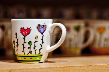 Foto de Una taza de cerámica blanca adornada con flores en forma de corazón y tallos verdes. Otras tazas similares son visibles en el fondo. - Imagen libre de derechos