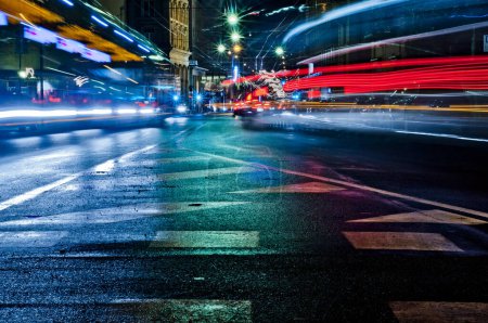 Foto de Una bulliciosa calle de la ciudad por la noche, capturada en una larga exposición. Las luces del vehículo atraviesan el asfalto mojado, iluminando los cruces peatonales. Energía urbana en movimiento. - Imagen libre de derechos