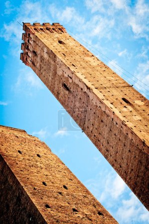 A low-angle view of a tall, narrow medieval stone tower with a textured surface, set against a clear blue sky with wispy clouds. Bologna. Italy
