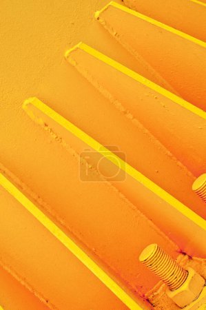 Foto de Vista de cerca de tres grandes pernos anclados en la superficie de hormigón amarillo, con soportes metálicos pintados del mismo color. Marco monocromático, fondo liso. - Imagen libre de derechos