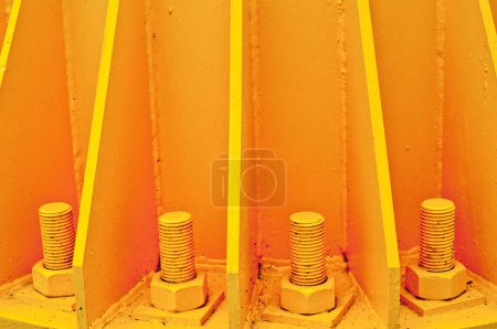 Foto de Vista de cerca de tres grandes pernos anclados en la superficie de hormigón amarillo, con soportes metálicos pintados del mismo color. Marco monocromático, fondo liso. - Imagen libre de derechos