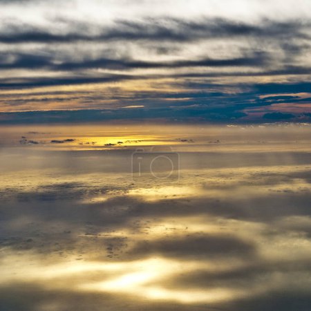 Luftaufnahme einer Wolkenlandschaft in der Abenddämmerung, die Wolkenschichten zeigt, wobei die untergehende Sonne einen sanften Schein wirft, die Ränder beleuchtet und Schatten erzeugt.