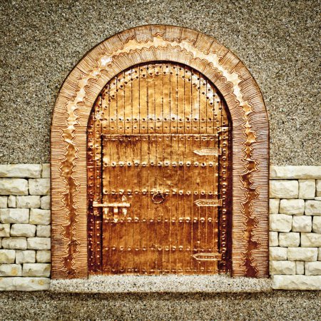A detailed wooden arched door adorned with intricate carvings and metal studs, set in a stone wall with a rough texture.
