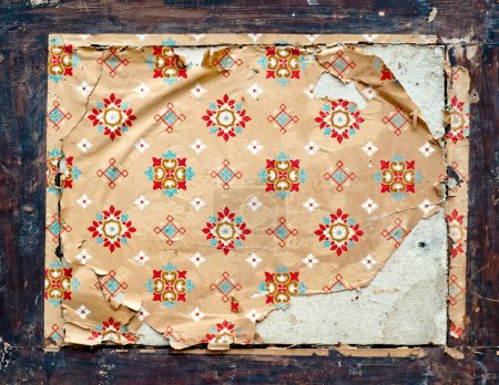A piece of old, patterned wallpaper with signs of wear and tear, adhered to a dark wooden surface. The wallpaper has a repetitive design featuring red and green motifs.