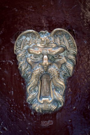Close-up photo of a decorative metal door knocker on a weathered wooden surface. Intricate design with scrollwork, lion head motif, and worn brass finish. Slight shadows and dust visible for a vintage feel.