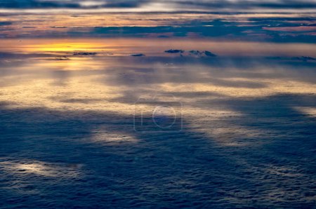 Vue aérienne d'un paysage nuageux au crépuscule, présentant des couches de nuages avec le coucher du soleil projetant une douce lueur, éclairant les bords et créant des ombres.