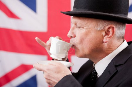 Traditioneller britischer Büroangestellter, der Tee trinkt, trägt dunklen Businessanzug mit passender Melone vor dem Hintergrund der Union-Jack-Flagge.