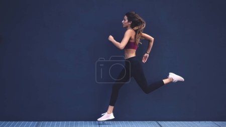 Foto de Joven mujer atlética caucásica retrato corriendo al aire libre contra la pared oscura. Estilo de vida activo. - Imagen libre de derechos