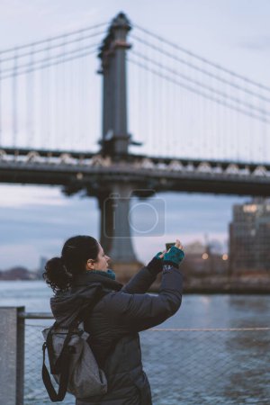 Photo for Woman taking pictures at Dumbo park in Brooklyn with Manhattan Bridge panoramic skyline view at sunset. New York City. - Royalty Free Image