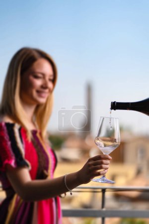 Téléchargez les photos : Verser du vin à une jeune femme à l'extérieur sur la terrasse de l'hôtel. Vue panoramique de la ville en arrière-plan. Concentre-toi sur le verre. Style de vie. - en image libre de droit