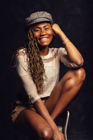 Téléchargez les photos : Belle femme noire africaine souriante assise sur un tabouret en bois portrait studio sur fond sombre. - en image libre de droit