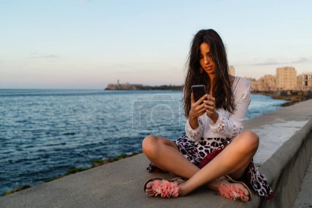 Photo for Beautiful Cuban woman portrait on the Malecon using smartphone. Havana, Cuba. Real life style image at sunset. - Royalty Free Image