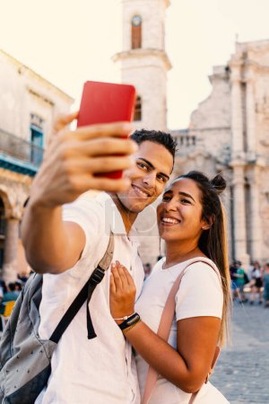Photo for Young smiling couple taking selfie with smartphone in Havana, Cuba. Cathedral in the background. Focus on the couple. - Royalty Free Image
