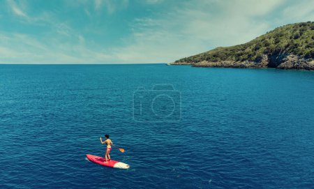 Téléchargez les photos : Femme d'âge moyen sur Stand Up Paddle Board, SUP, par une journée ensoleillée en été. Vue aérienne. Île de Giannutri, Italie. Concept de vie active. Image filtrée. - en image libre de droit