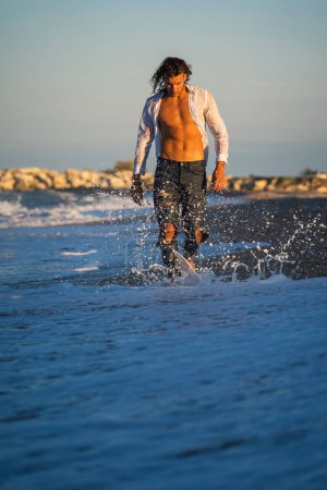 Téléchargez les photos : Youn fit homme sexy marchant pieds nus sur la plage au coucher du soleil portant jeans et chemise. - en image libre de droit