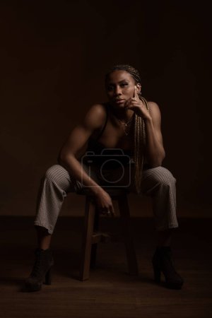 Foto de Retrato seguro de mujer negra africana sentada en taburete de madera sobre fondo oscuro. - Imagen libre de derechos