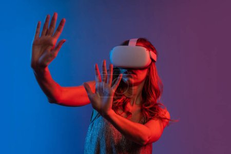 Foto de Mujer joven con auriculares para realidad aumentada - gafas en realidad virtual. Chica manos arriba con gafas de realidad virtual y tocar el aire durante la experiencia VR. Retrato de estudio con geles de color. - Imagen libre de derechos