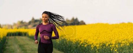 Téléchargez les photos : Portrait de jeune femme sportive courant à l'extérieur sur la campagne au coucher du soleil dans un champ jaune. Image panoramique. - en image libre de droit