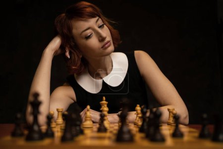 Foto de Hermosa mujer pelirroja jugando ajedrez sobre fondo oscuro. Antiguo efecto de película añadido en la postproducción. - Imagen libre de derechos