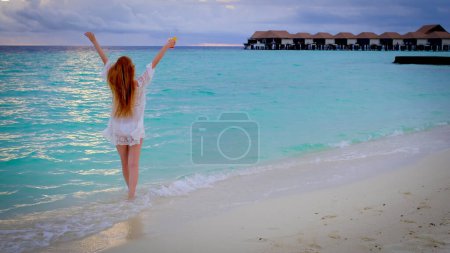 Téléchargez les photos : Femme avec un cocktail au coucher du soleil aux Maldives. La fille est heureuse et lève la main. Océan bleu turquoise sur le fond. Voyage d'été concept de vacances. - en image libre de droit