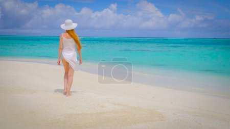 Photo for Woman in a bikini is walking with a white hat. Female is happy on Maldives. Blue turquoise ocean on the background. Girl enjoys her tropical holidays. Summer travel vacation concept. - Royalty Free Image