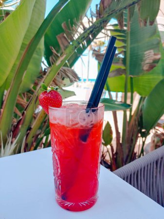 Téléchargez les photos : Un cocktail rouge avec une fraise est debout sur une table. Des paumes vertes sur le fond. Concept de fête de vacances. Détendez-vous fond froid. - en image libre de droit