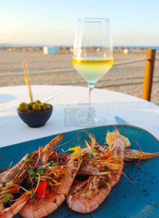 Téléchargez les photos : Gros plan de crevettes grillées sur une assiette avec un verre de vin et d'olives. La plage et la mer sont en arrière-plan. La nourriture méditerranéenne est sur la table. - en image libre de droit