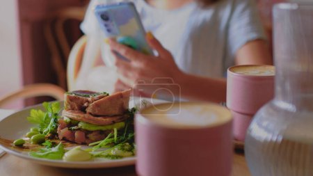 Photo for Coffee and a breakfast is on the table. Avocado and green veggie food. Tuna meat. Women is with a mobile phone on a background. Blogger concept. - Royalty Free Image