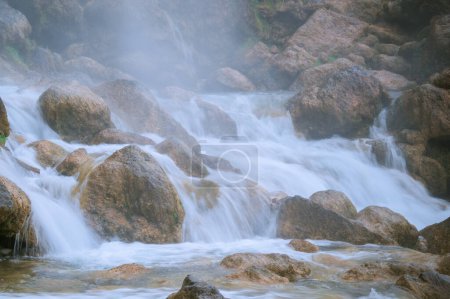 The image captures a close-up of water flowing over large rocks, creating a series of small cascades and gentle rapids. Mist rises from the water, adding a serene and ethereal atmosphere to the scene.