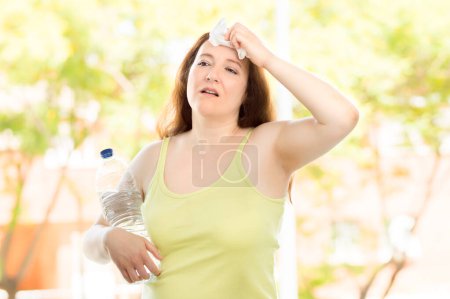Foto de una mujer infeliz sudando sufriendo un golpe de calor en el parque con una botella de agua
