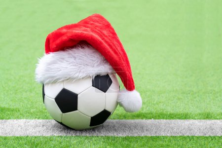 Téléchargez les photos : Gros plan du chapeau rouge du Père Noël sur le ballon de football à l'herbe verte au stade avec espace de copie. - en image libre de droit