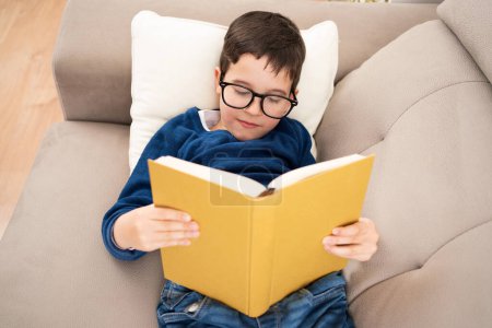 Photo for Above view portrait of cute boy wearing big glasses reading book while lying on couch, homeschooling concept and back to school and copy space - Royalty Free Image
