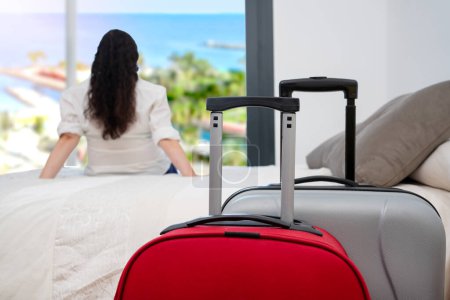 Rückansicht einer einzelnen Touristin, die nach ihrer Ankunft mit Koffern im Vordergrund entspannt aus einem Hotelzimmerfenster blickt