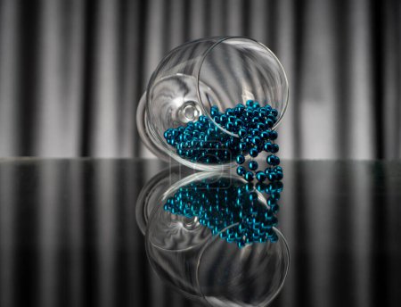 Téléchargez les photos : Perles brillantes et turquoise suspendues à un verre couché sur le côté sur une surface réfléchissante - en image libre de droit
