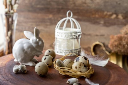 Téléchargez les photos : Œufs de caille dans un nid couché sur une surface en bois sombre, sur le fond d'un lapin de Pâques en porcelaine blanche et une cage avec des plumes - en image libre de droit