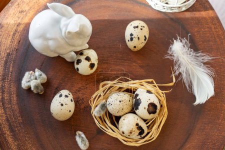 Téléchargez les photos : Œufs de caille dans un nid couché sur une surface en bois sombre, sur le fond d'un lapin de Pâques en porcelaine blanche - en image libre de droit